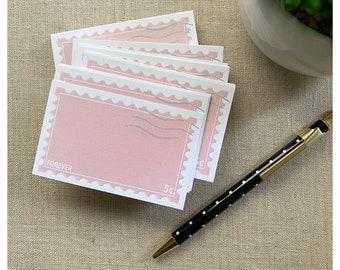 May include: A stack of pink notecards with a postage stamp design. Each card has a white border and the words "FOREVER" and "5¢" printed on it. A black and white polka dot pen with gold accents rests on the surface.