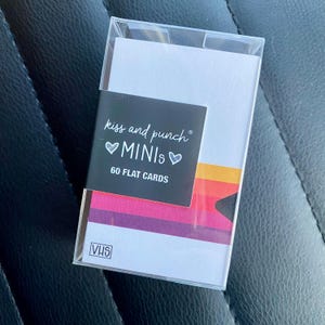 Puede incluir: Una caja de plástico transparente que contiene 60 tarjetas planas con una etiqueta negra que dice kiss and punch MINIs 60 FLAT CARDS. Las tarjetas presentan un diseño de rayas horizontales en naranja, rojo y morado, con un logo VHS abajo.