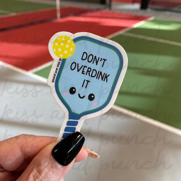 May include: A sticker featuring a blue pickleball paddle with the text "DON'T OVERDINK IT." The paddle has a smiling face and a yellow pickleball attached. The sticker is held in a hand.