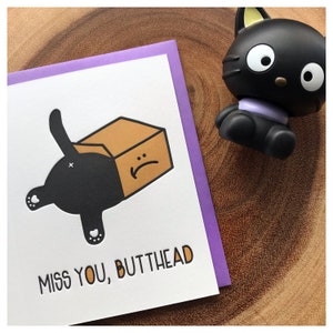 May include: A greeting card with a black cat illustration, featuring a cat's rear end sticking out of a cardboard box. The text "MISS YOU, BUTTHEAD" is printed below the illustration. A black cat figurine is next to the card.