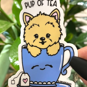 Może przedstawiać: Biała naklejka z kreskówkową ilustracją małego psa siedzącego w niebieskiej filiżance do herbaty. Tekst "Pup of Tea" jest nadrukowany nad psem. Filiżanka do herbaty ma uśmiechniętą twarz, a z boku zwisa torebka herbaty z różowym sercem.