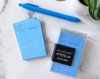 May include: A light blue pen rests near two stacks of blue notecards with lines and headings. A clear plastic case holds a stack of blue cards with a black label that reads "kiss and punch MINIs 60 FLAT CARDS."
