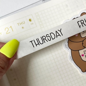 May include: A strip of white washi tape with the word THURSDAY printed in black sans-serif font, positioned over a grid-patterned planner page that displays the number 21, the text THU, and a moon icon, next to a brown bear sticker.