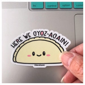 May include: White sticker with a cartoon illustration of a smiling yellow gyoza with the text "Here we gyoz-again!" in black lettering. The sticker has a pink heart on the gyoza and the text "Kiss and Punch" in small black lettering at the bottom.