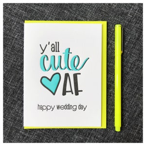 May include: A white greeting card with a neon yellow border. The card features the text "y'all cute AF" in black and teal lettering, with a teal heart. The words "happy wedding day" are printed below. A neon yellow pen is next to the card.