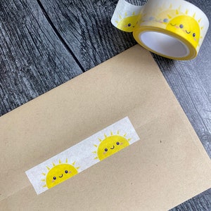 May include: A roll of clear washi tape with a repeating pattern of yellow smiling suns. The tape is on a brown paper surface.