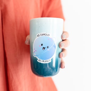 May include: A blue and green ombre mug with a sticker of a cartoon planet with a face and the text "As Famous As Uranus".