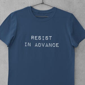 Puede incluir: Una camiseta azul marino con el texto blanco "RESIST IN ADVANCE" impreso en el pecho. La camiseta está colgada de una percha negra sobre un fondo gris. La camiseta está hecha de un material suave.
