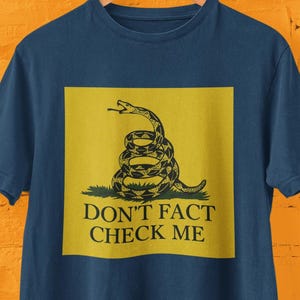 Könnte beinhalten: Ein marineblaues T-Shirt mit einem gelben Quadrat, auf dem sich eine gewickelte Schlange mit dem Text "Don't Fact Check Me" befindet.