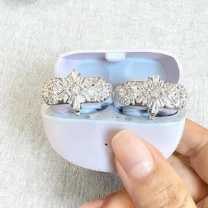 Puede incluir: Un par de pendientes plateados con diseño de estrella, dentro de un estuche lila claro. Los pendientes son muy detallados con múltiples facetas, que reflejan la luz. El estuche es sostenido por una mano.