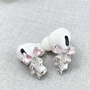 Puede incluir: Auriculares inalámbricos blancos con adornos de lazo rosa en forma de corazón. Los lazos son plateados con un dije de estrella colgante. Los auriculares están sobre una superficie gris texturizada. Accesorio decorativo para auriculares.