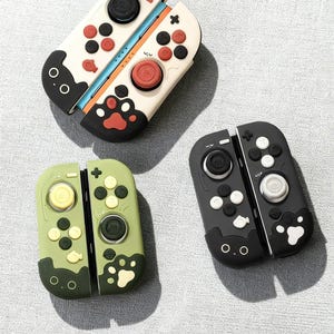 May include: Three colourful game controller accessories. One is white with red and black buttons and a paw print design. Another is olive green with yellow and green buttons. The third is black with white buttons and a fish design.