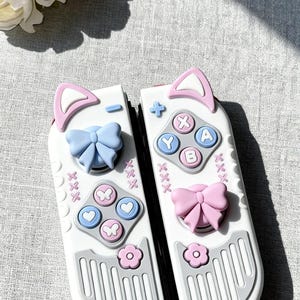 May include: White and grey game controller accessories with pink cat ear accents, blue and pink bows, and colourful buttons. The controllers have a cute, playful design, ideal for gamers.