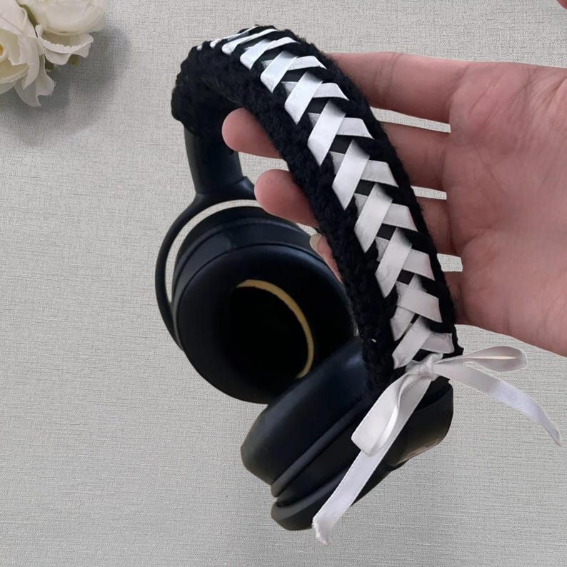May include: Black headphones with a braided headband featuring white ribbon accents. The headband is a crocheted design. A white ribbon is tied at the base of the headband. The headphones are black with a white accent.