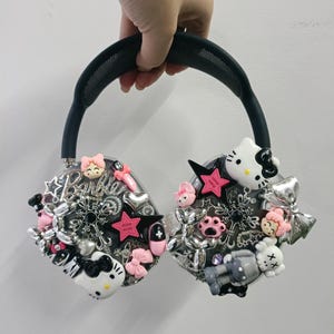 K&ouml;nnte beinhalten: Individuell gestalteter schwarzer Kopfh&ouml;rer, verziert mit verschiedenen Charms, darunter rosa Sterne, Schleifen und ein Hello Kitty-Design. Die Ohrmuscheln tragen das Wort "Barbie" und andere dekorative Elemente. Das Kopfband ist dunkelgr&uuml;n.