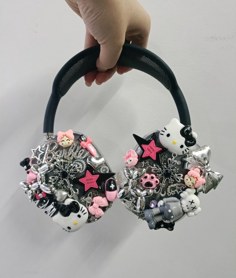 K&ouml;nnte beinhalten: Individuell gestalteter schwarzer Kopfh&ouml;rer, verziert mit verschiedenen Charms, darunter rosa Sterne, Schleifen und ein Hello Kitty-Design. Die Ohrmuscheln tragen das Wort "Barbie" und andere dekorative Elemente. Das Kopfband ist dunkelgr&uuml;n.