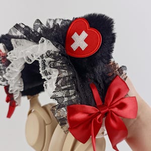 May include: Headphones with a black fur and lace headband, adorned with a red heart-shaped patch featuring a white cross. A large red satin bow and smaller red accents add to the design. The headphones are beige.