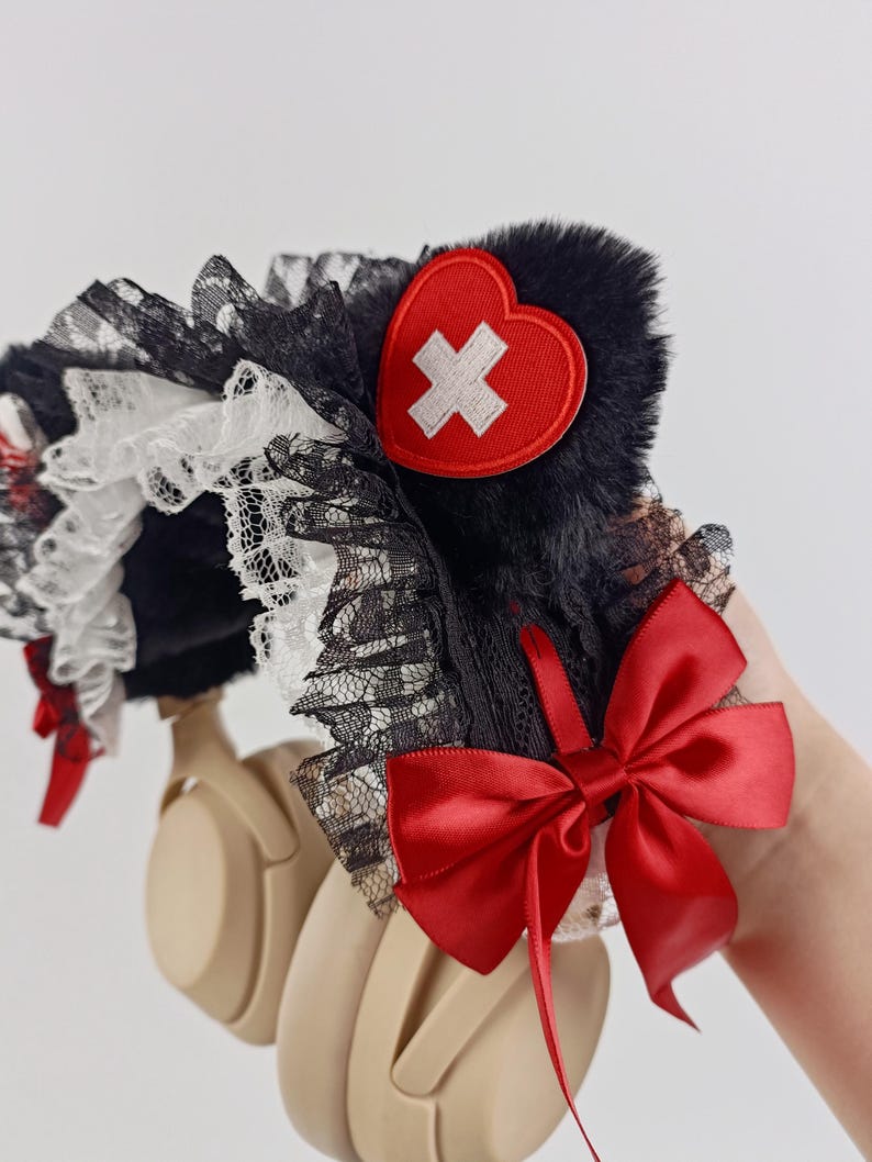 May include: Headphones with a black fur and lace headband, adorned with a red heart-shaped patch featuring a white cross. A large red satin bow and smaller red accents add to the design. The headphones are beige.