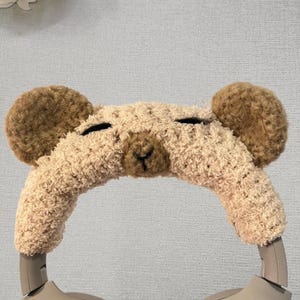 Könnte beinhalten: Eine beige Teddybär-Kopfhörerhülle mit braunen Ohren und schwarz gestickten Augen und Nase. Die Hülle ist so konzipiert, dass sie über das Kopfband eines Kopfhörers passt und eine dekorative und verspielte Note verleiht.