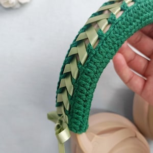 May include: A green crocheted headband with woven light green ribbons. The headband is shown over a pair of beige headphones. A light green ribbon is tied in a bow on the side of the headband. The background is a light blue.