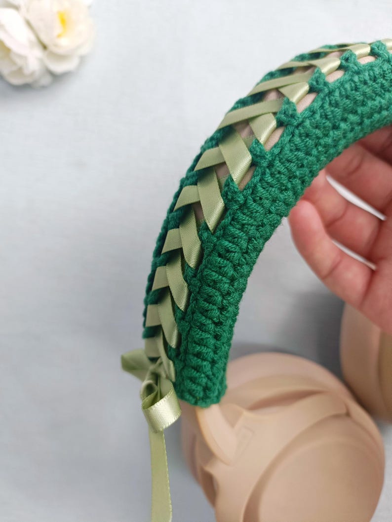 May include: A green crocheted headband with woven light green ribbons. The headband is shown over a pair of beige headphones. A light green ribbon is tied in a bow on the side of the headband. The background is a light blue.