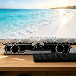 Puede incluir: Un monitor de computadora con un borde de encaje blanco con pequeños lazos a lo largo del borde inferior y detalles en forma de alas blancas en las esquinas. Debajo del monitor hay una barra de sonido negra y blanca con alas laterales.