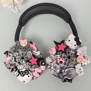 K&ouml;nnte beinhalten: Schwarze Over-Ear-Kopfh&ouml;rer, verziert mit verschiedenen Charms. Die Charms umfassen rosa Sterne, Hello Kitty-Figuren, Schleifen und andere dekorative Elemente. Die Kopfh&ouml;rer haben ein schwarzes Kopfband und Ohrmuscheln, vor einem hellblauen Hintergrund.