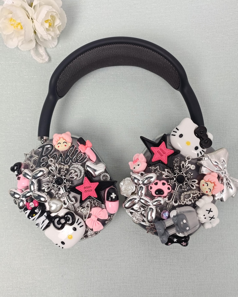 K&ouml;nnte beinhalten: Schwarze Over-Ear-Kopfh&ouml;rer, verziert mit verschiedenen Charms. Die Charms umfassen rosa Sterne, Hello Kitty-Figuren, Schleifen und andere dekorative Elemente. Die Kopfh&ouml;rer haben ein schwarzes Kopfband und Ohrmuscheln, vor einem hellblauen Hintergrund.