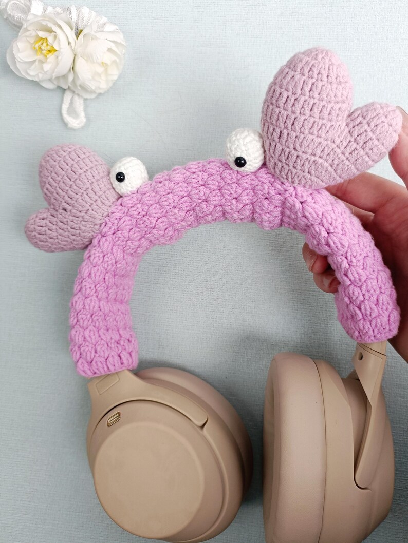 Puede incluir: Fundas para auriculares de crochet rosa en forma de cangrejo con ojos blancos. Las fundas est&aacute;n en un par de auriculares beige.