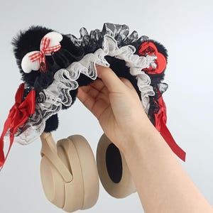 May include: A black, white, and red decorative headphone accessory. The accessory features black faux fur, white lace trim, and red ribbon accents. A hand is holding the headphones.