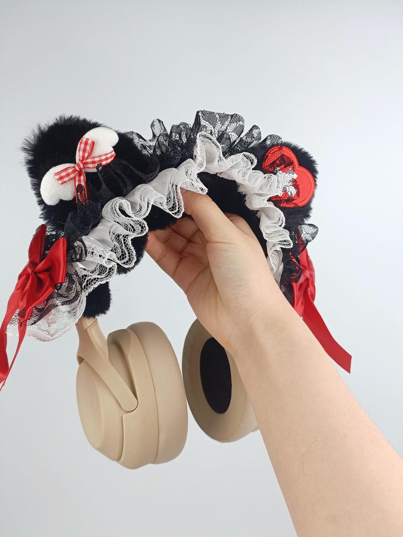 May include: A black, white, and red decorative headphone accessory. The accessory features black faux fur, white lace trim, and red ribbon accents. A hand is holding the headphones.