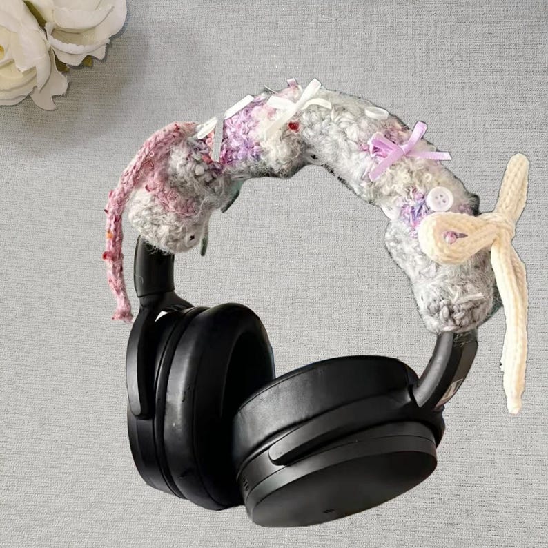 May include: Black over-ear headphones with a crocheted headband cover. The cover is primarily gray with pink and purple accents, small white bows, and a cream-colored bow on the side. The headphones are set against a neutral background.