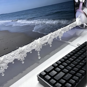 Peut inclure: Une bordure de moniteur en dentelle blanche ornée de motifs floraux complexes, de petits nœuds en tissu et de perles transparentes suspendues. Cette garniture décorative est fixée au bord inférieur d'un écran d'ordinateur, au-dessus d'un clavier noir.