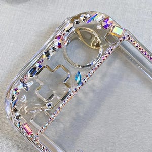 May include: A clear phone case embellished with iridescent rhinestones and crystals. The case has a cross-shaped design and a border of small, colourful gems. The transparent design allows the phone's colour to show through, creating a sparkling effect.
