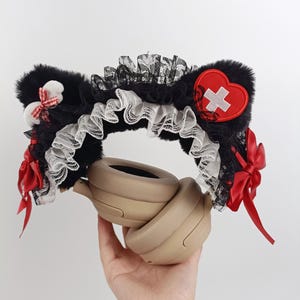 May include: Black faux fur cat ear headband with white lace trim, red bows, and a red heart patch with a white cross. The headband is designed to be worn with headphones.