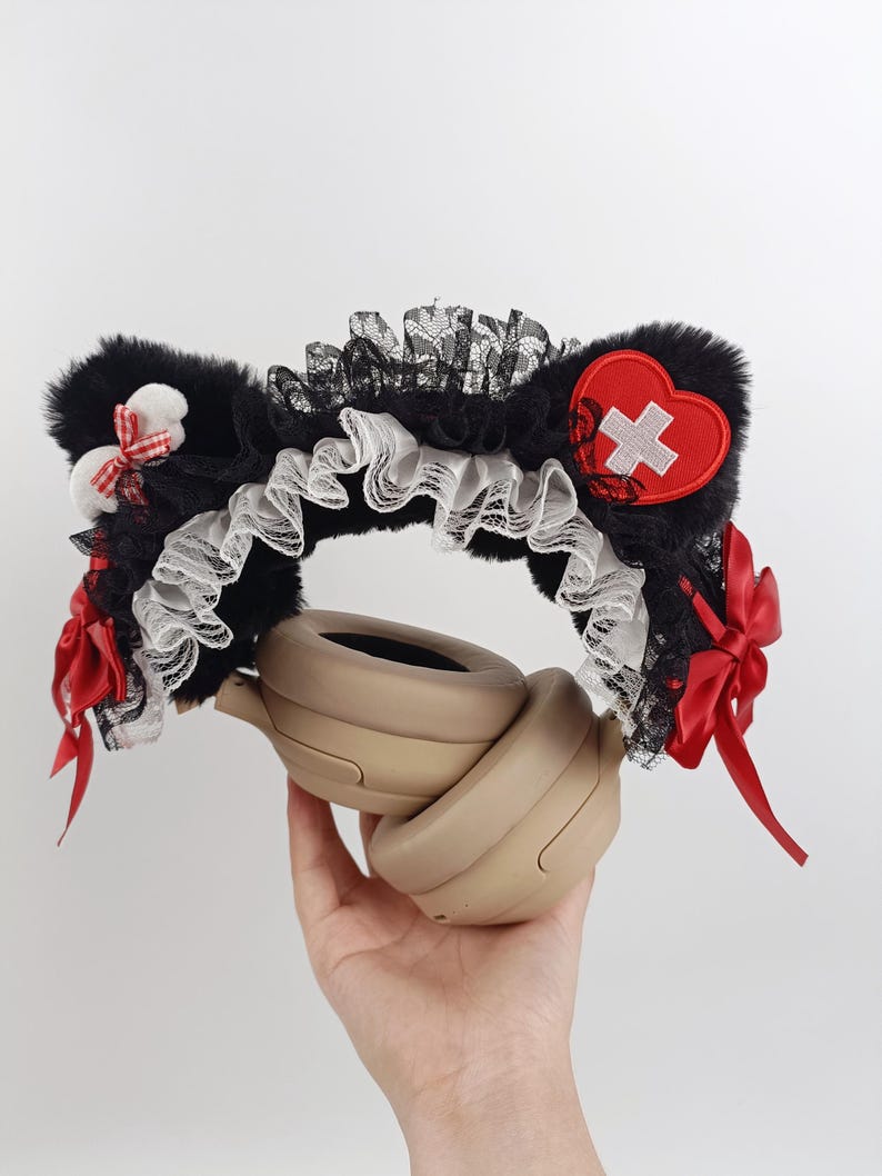 May include: Black faux fur cat ear headband with white lace trim, red bows, and a red heart patch with a white cross. The headband is designed to be worn with headphones.