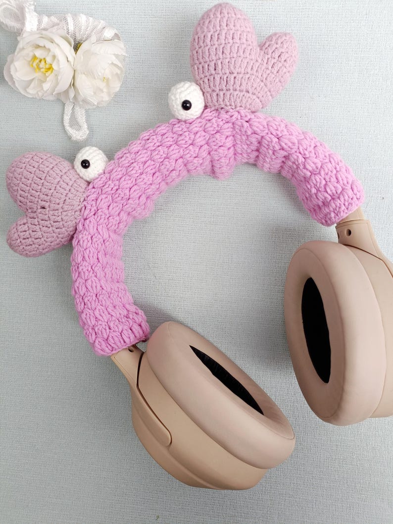 Puede incluir: Funda de auriculares de ganchillo rosa con ojos saltones y parte superior en forma de coraz&oacute;n. La funda est&aacute; en unos auriculares beige.