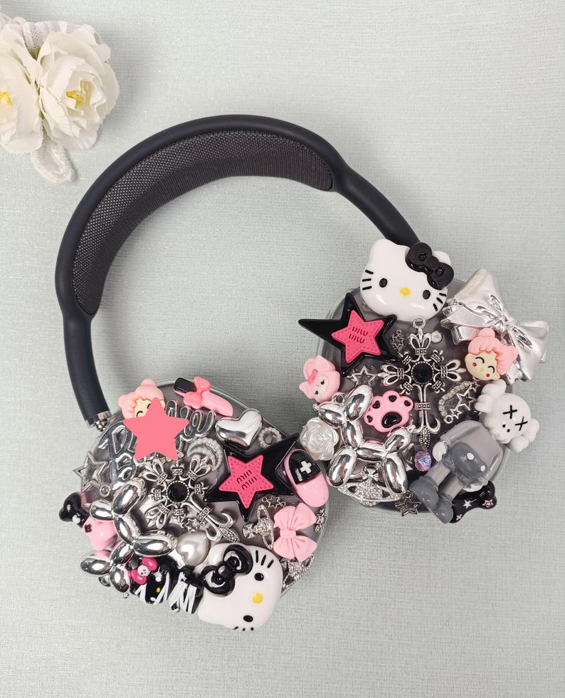 K&ouml;nnte beinhalten: Schwarzer Over-Ear-Kopfh&ouml;rer, verziert mit verschiedenen Charms, darunter rosa Sterne, Schleifen und eine Hello Kitty-Figur. Der Kopfh&ouml;rer hat ein schwarzes Kopfband und Ohrmuscheln mit Mesh-Struktur.