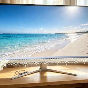 Könnte beinhalten: Ein Computermonitor mit einem dünnen schwarzen Rahmen, der an der Unterkante mit weißer Spitzenborte verziert ist. Die Spitze ist mit kleinen Schleifen und hängenden Perlen geschmückt. Ein großes weißes Blumenornament ist links angebracht.