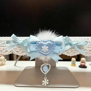 Peut inclure: Un nœud en satin bleu clair avec un pompon en fausse fourrure blanche, orné d'un cœur central avec un flocon de neige blanc, et une breloque argentée avec une perle bleu clair et un petit pendentif flocon de neige suspendu au nœud.