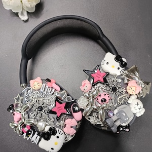 K&ouml;nnte beinhalten: Schwarzer Kopfh&ouml;rer, verziert mit verschiedenen Charms, darunter Hello Kitty, Sterne, Schleifen und das Wort "Barbie". Die Charms sind in Pink, Silber und Schwarz gehalten. Der Kopfh&ouml;rer hat ein schwarzes Kopfband und Ohrmuscheln.