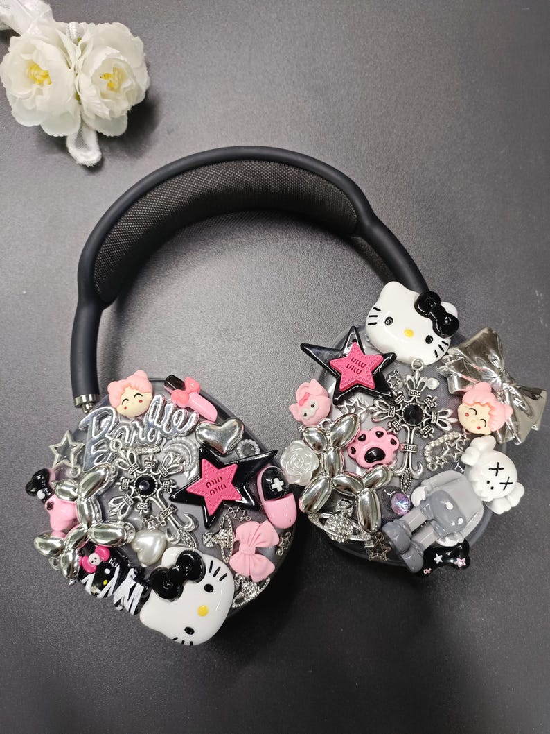 K&ouml;nnte beinhalten: Schwarzer Kopfh&ouml;rer, verziert mit verschiedenen Charms, darunter Hello Kitty, Sterne, Schleifen und das Wort "Barbie". Die Charms sind in Pink, Silber und Schwarz gehalten. Der Kopfh&ouml;rer hat ein schwarzes Kopfband und Ohrmuscheln.
