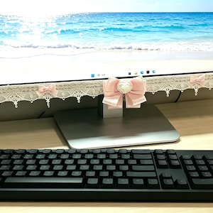 Puede incluir: Un marco de monitor de computadora decorado con ribete de encaje blanco y pequeños lazos de tela rosa. Un lazo rosa más grande con un adorno central en forma de corazón está sujeto al centro del borde inferior. Un teclado negro está en primer plano.