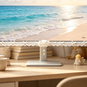 May include: A computer monitor frame decorated with white lace trim featuring small bows along the edge and a central white bow ornament with a dangling beaded charm. The screen displays a bright, sunny beach scene with blue ocean waves.