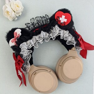 May include: Black faux fur headphone accessory with red and white accents. Features a red heart with a white cross, red bows, and white lace trim. The headphones are a neutral beige color.