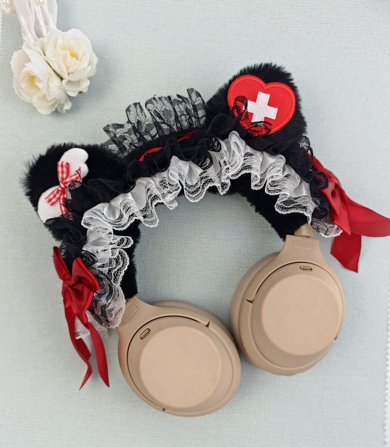 May include: Black faux fur headphone accessory with red and white accents. Features a red heart with a white cross, red bows, and white lace trim. The headphones are a neutral beige color.