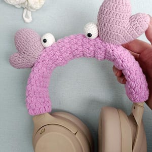 Puede incluir: Fundas para auriculares de crochet rosa con ojos saltones y orejas en forma de coraz&oacute;n. Las fundas est&aacute;n en un par de auriculares beige.