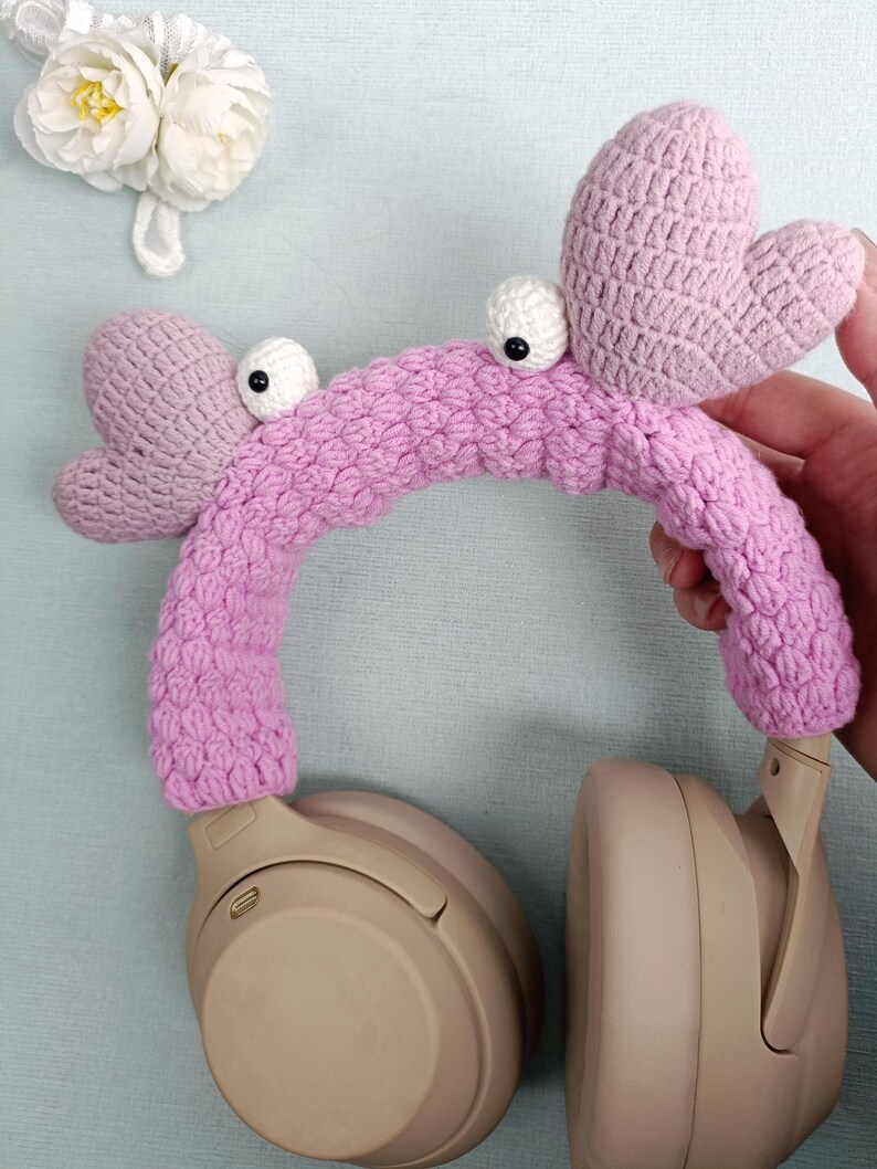 Puede incluir: Fundas para auriculares de crochet rosa con ojos saltones y orejas en forma de coraz&oacute;n. Las fundas est&aacute;n en un par de auriculares beige.