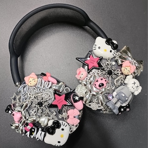 K&ouml;nnte beinhalten: Schwarzer Kopfh&ouml;rer mit verschiedenen Anh&auml;ngern, darunter Hello Kitty, Sterne, Schleifen und eine Roboterfigur. Die Charms sind in Rosa, Silber und Wei&szlig; gehalten und schaffen ein verspieltes und dekoratives Design.