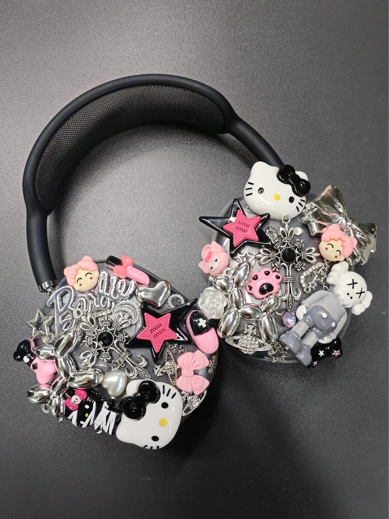 K&ouml;nnte beinhalten: Schwarzer Kopfh&ouml;rer mit verschiedenen Anh&auml;ngern, darunter Hello Kitty, Sterne, Schleifen und eine Roboterfigur. Die Charms sind in Rosa, Silber und Wei&szlig; gehalten und schaffen ein verspieltes und dekoratives Design.